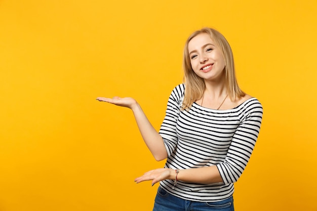 Portrait of smiling attractive young woman in striped clothes pointing hands aside isolated on yellow orange wall background in studio. People sincere emotions, lifestyle concept. Mock up copy space.