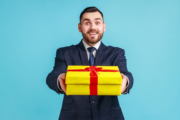 Portrait of smiling bearded businessman wearing dark official style suit giving wrapped gift box and smiling at camera happy holiday Indoor studio shot isolated on blue background