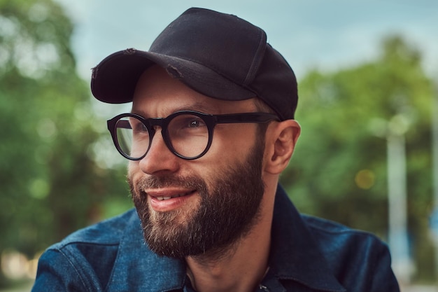 Ritratto di un hipster barbuto sorridente in occhiali e berretto all'aperto.