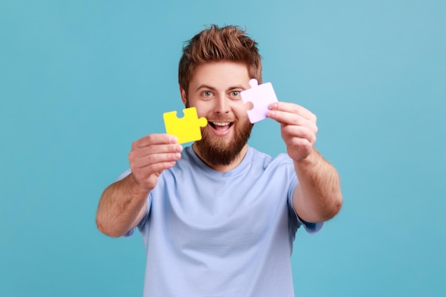 Portrait of smiling bearded man holding two puzzle parts and smiling joyfully ready to connect jigsaw pieces symbol of union and association Indoor studio shot isolated on blue background