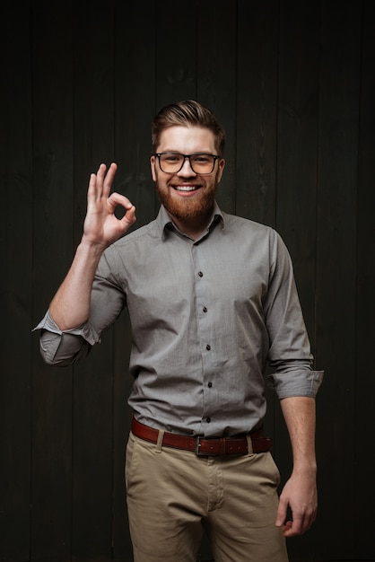 Portrait of a smiling bearded man wearing eyeglasses and shirt and showing okay gesture isolated on the black wooden surface
