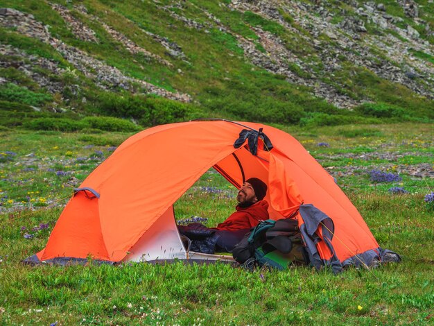 텐트에 앉아 웃는 수염된 관광객의 초상화. 솔로 트레킹. 여름의 녹색 바위가 많은 고지대에서 캠핑. 자연의 평화와 휴식.