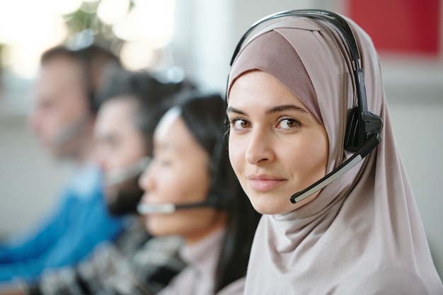 Portrait of smiling beautiful Arabian contact center agent in hijab using wireless headset with microphone while answering customers