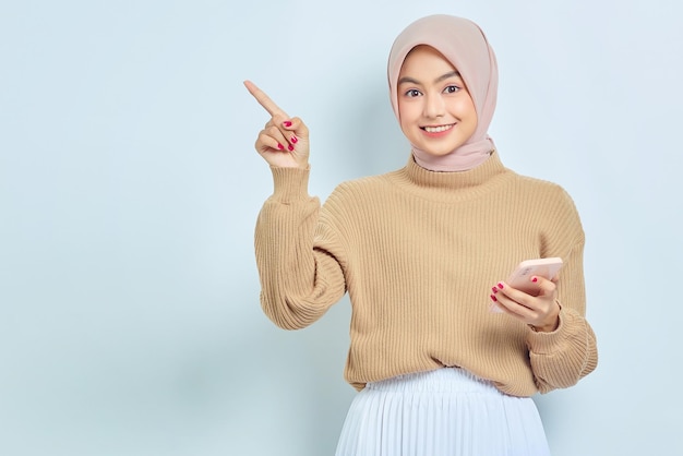 Photo portrait of smiling beautiful asian muslim woman in brown sweater and hijab holding mobile phone and pointing finger aside isolated over white background people religious lifestyle concept