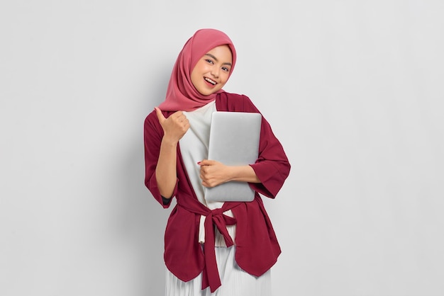 Portrait of smiling beautiful asian woman in casual shirt and\
hijab holding a laptop, showing thumb up isolated over white\
background. people religious lifestyle concept