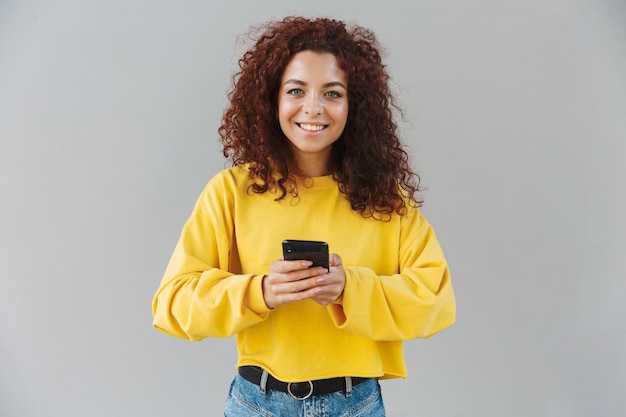 Portrait of smiling beautiful curly woman isolated over gray wall using mobile phone.