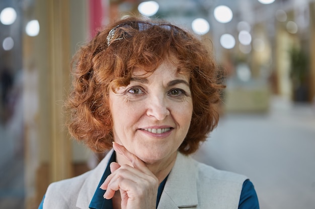 Portrait of smiling beautiful old lady with red curly short hair in shopping mall.