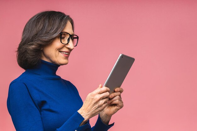 Portrait of smiling beautiful senior aged mature woman using tablet computer, isolated over pink background. Copy space for text.