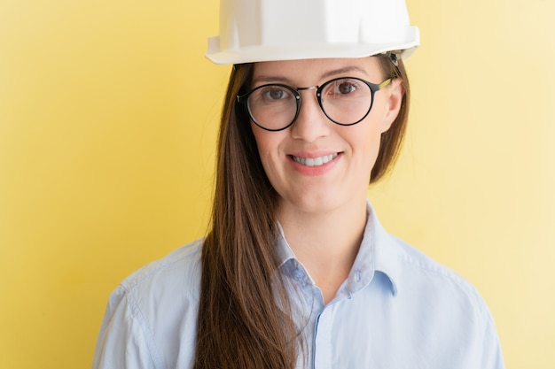 黄色の背景に分離された建設ヘルメットとメガネで笑顔の美しいタタール人女性エンジニアの肖像画