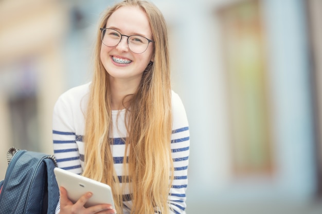 歯のブレースと笑顔の美しい10代の少女の肖像画。ランドセルとタブレットデバイスを持つ若い女子高生。