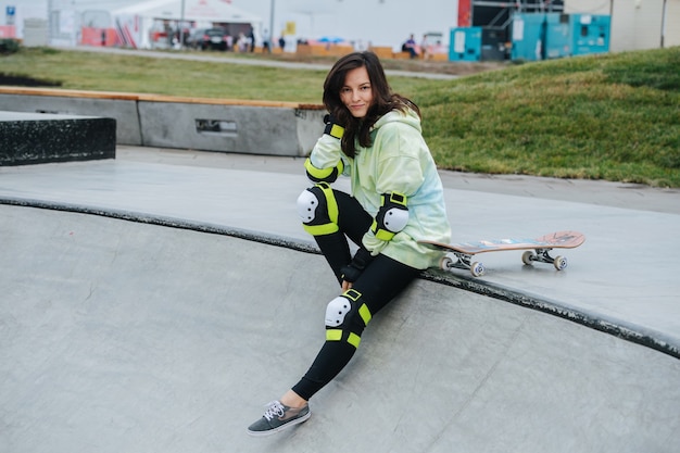 Portrait of a smiling beautiful woman skater on a deck. She wears some protective gear. She's leaning on her hand, looking at the camera.