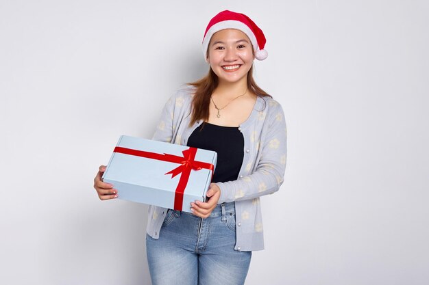 Portrait of a smiling beautiful young asian woman wearing a\
blue sweater with a santa claus hat holding a gift box isolated on\
white studio background merry christmas concept