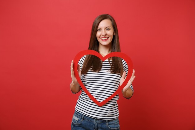 Portrait of smiling beautiful young woman in casual striped clothes holding big red wooden heart isolated on bright red wall background. People sincere emotions, lifestyle concept. Mock up copy space.