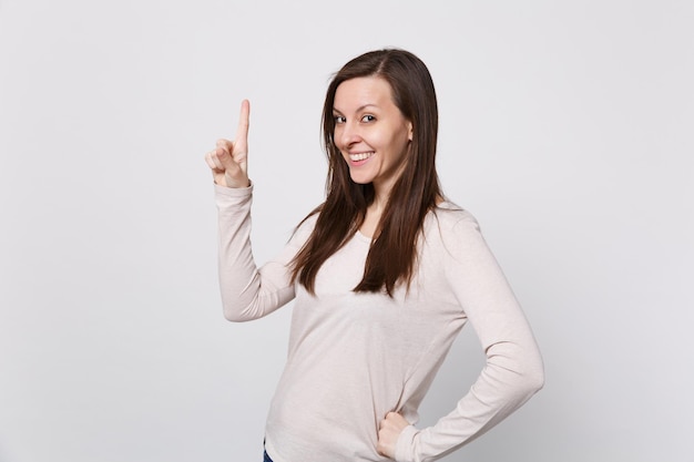 Portrait of smiling beautiful young woman in light clothes standing pointing index finger up isolated on white wall background in studio. People sincere emotions lifestyle concept. Mock up copy space.