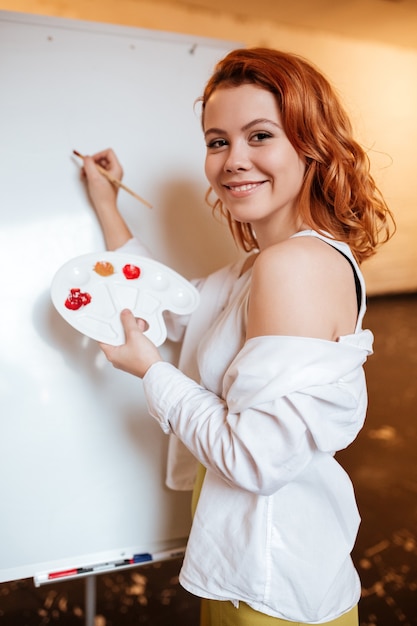 예술가 워크숍에서 빈 캔버스에 오일 페인트로 붉은 머리 그림을 그린 웃고 있는 아름다운 젊은 여성 화가의 초상화