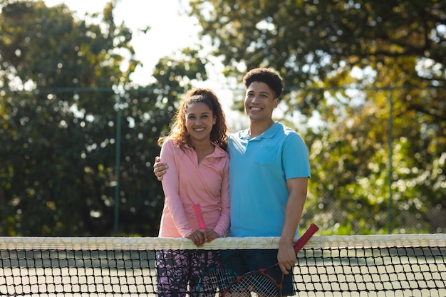 Portrait of smiling biracial couple playing tennis embracing on sunny outdoor tennis court. Inclusivity, sport, healthy hobbies, competition, fitness and leisure time concept.