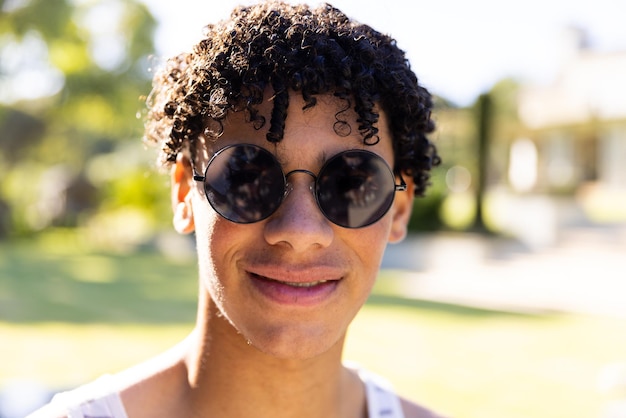 Portrait of smiling biracial fit man wearing sunglasses and vest standing in sunny garden. Summer, free time, relaxation and vacations.