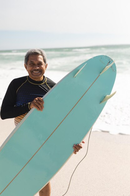 Portrait of smiling biracial senior man holding blue surfboard at beach against sky during sunny day