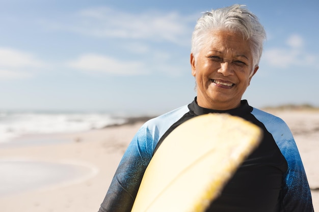 Portrait of smiling biracial senior woman carrying surfboard at beach against sky on sunny day