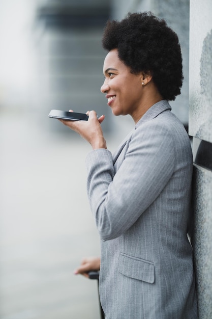 Portrait of a smiling black business woman talking voice\
message on a smart phone in front a corporate building.