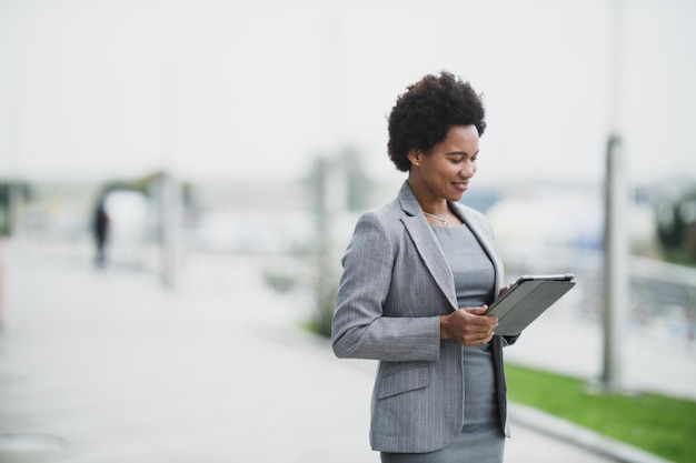 Portrait of a smiling black business woman using app on a\
digital tablet in front a corporate building.