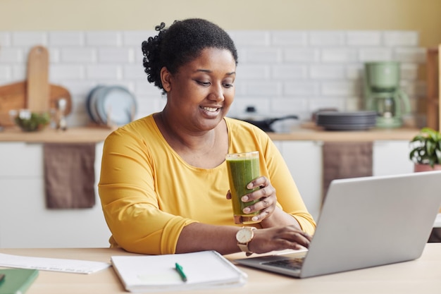 Ritratto di donna nera sorridente che beve frullato e usa il laptop a casa