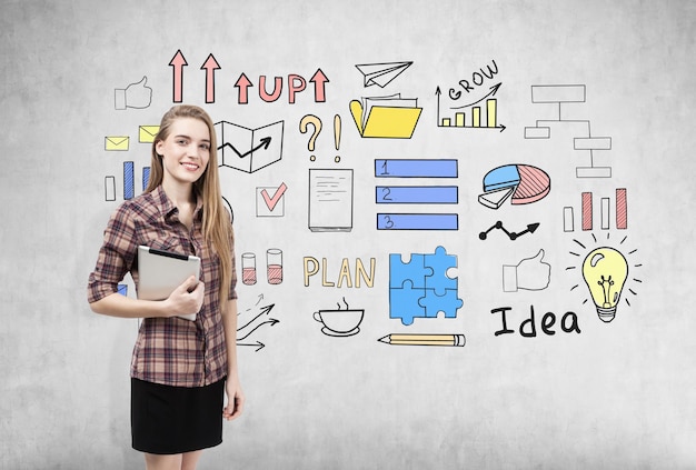 Photo portrait of a smiling blond woman in a checkered shirt standing with her tablet near a concrete wall with colorful business idea icons. concept of business education