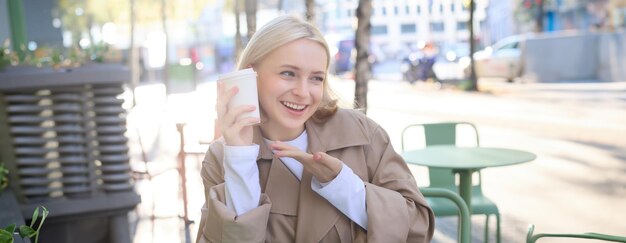 Foto ritratto di una donna bionda sorridente che indica con il dito la sua tazza di caffè.