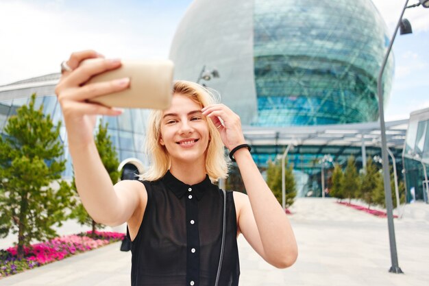 Portrait of a smiling blonde girl in black makes selfie photo on cellphone on the background of a glass business building