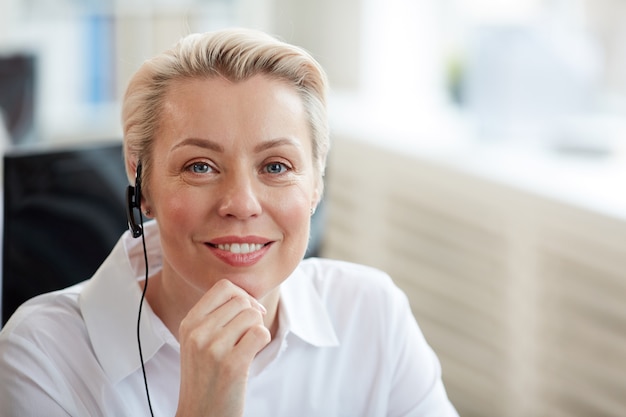 Portrait of smiling blonde woman wearing headset and looking while working in support service call center