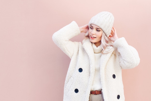 Portrait of a smiling blonde woman in winter clothes looking away on pink background.