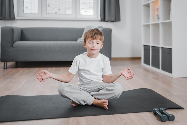 Portrait of smiling boy child on yoga meditation session, sitting in Padmasana exercise, Lotus pose with Namaste, working out wearing home clothes, eyes closed.
