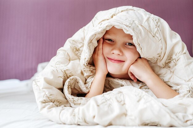 Photo portrait of smiling boy hiding under blanket in bed at home