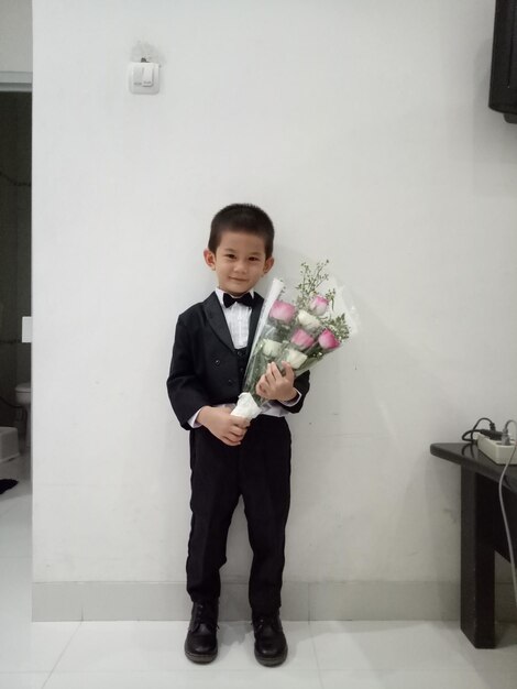 Photo portrait of smiling boy holding flower bouquet against white wall at home