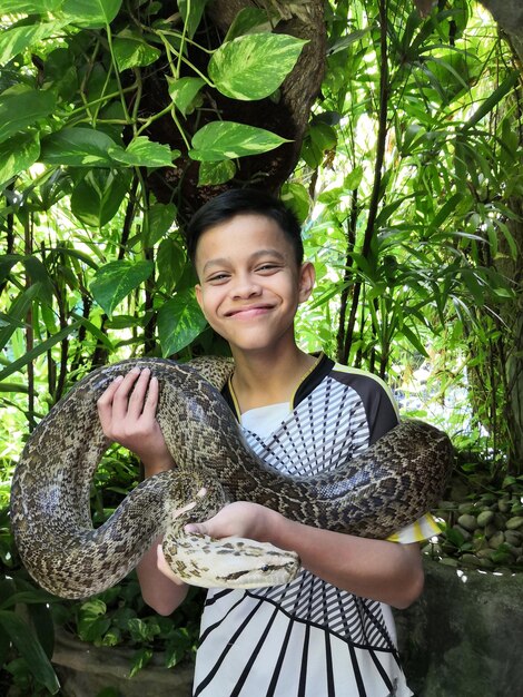 Foto ritratto di un ragazzo sorridente che tiene un serpente contro le piante