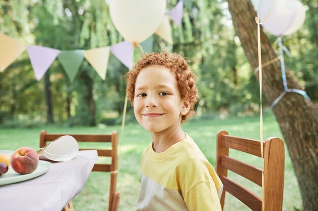 夏の誕生日パーティーを楽しんで屋外のピクニックテーブルでカメラを見ている笑顔の少年の肖像画