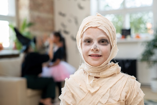 Portrait of smiling boy with white face wrapped up into bandages like mummy standing in room decorated for Halloween party