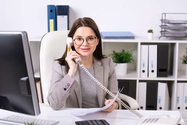 Portrait of smiling brunette secretary sitting at desk and answering phone call in office