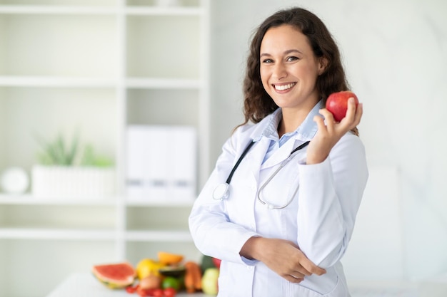 Foto ritratto di donna bruna sorridente che lavora in clinica tenendo mela