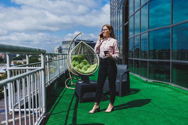 Portrait of smiling business woman reading sms on smartphone\
during coffee break on office balcony modern terrace in the office\
for relaxation
