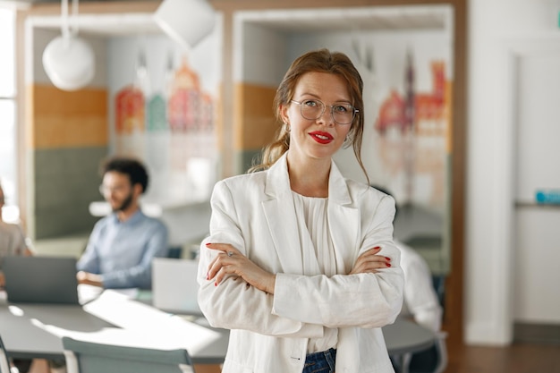 Portrait of smiling businesswoman in white suit standing in modern office on colleagues background