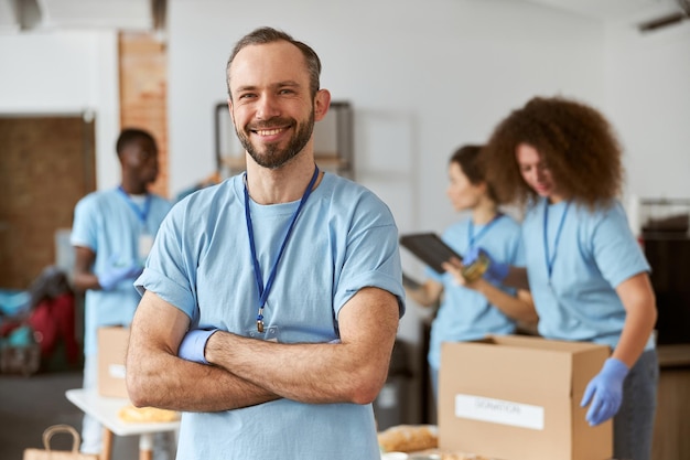 青い制服を着た笑顔の白人男性ボランティアの肖像画と腕を組んで立っている保護手袋 段ボール箱に商品を梱包するチーム