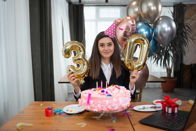 Ritratto di donna caucasica sorridente seduto al tavolo, torta di compleanno e numero 30 in webcam.