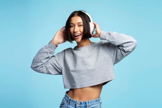 Photo portrait of smiling cheerful asian woman wearing stylish casual clothes listening to music in headphones looking at camera standing isolated on blue background concept of positive lifestyle