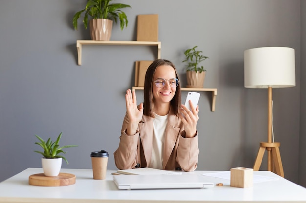 Portrait of smiling cheerful Caucasian woman sitting at workplace with smart phone in hands having video call with business partner waving hand saying hello