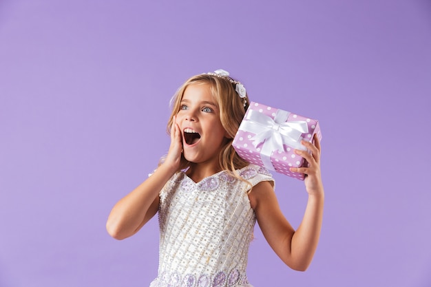 Portrait of a smiling cheerful pretty girl dressed in a princess dress isolated over violet wall, holding present box