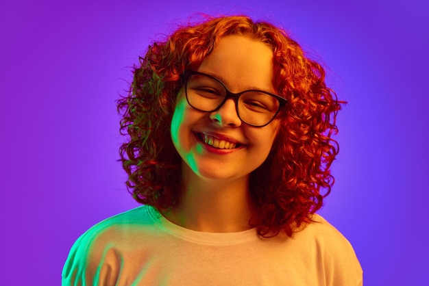 Portrait of smiling cheerful teen girl with curly redhead hair in glasses lookin at camera against