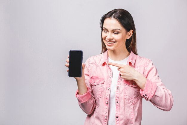 白い背景に分離された空白のスマートフォンの画面を示す笑顔の陽気な女性の肖像画