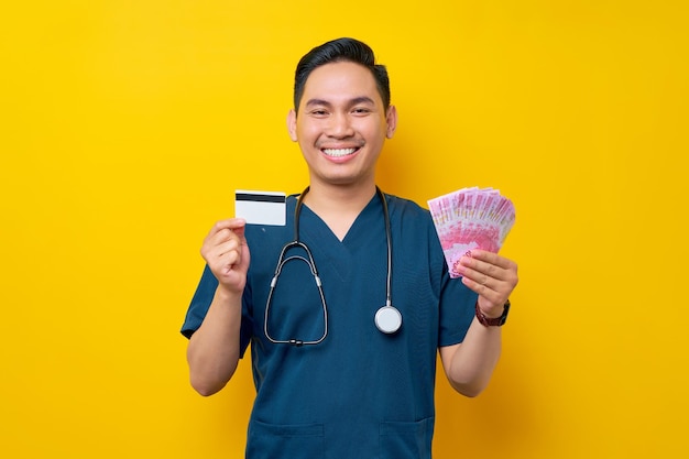 Portrait of a smiling cheerful young Asian male doctor or nurse wearing blue uniform holding cash money and credit card isolated on yellow background Healthcare medicine concept