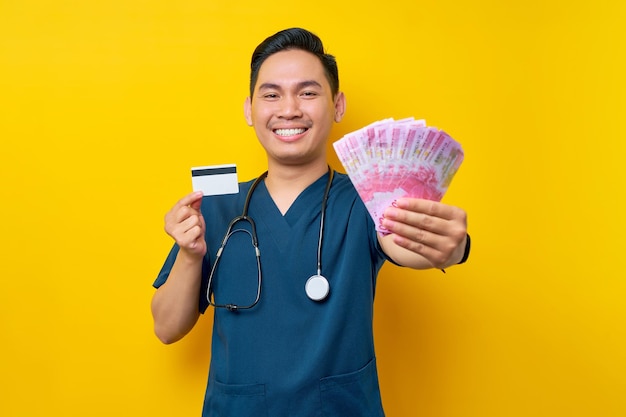 Portrait of a smiling cheerful young Asian male doctor or nurse wearing blue uniform holding cash money and credit card isolated on yellow background Healthcare medicine concept